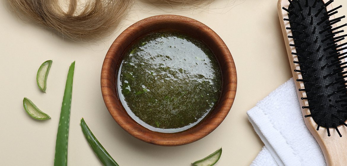 Aloe Vera, Schale mit grünem Haaröl, ein Handtuch, eine Bürste und ein brauner Haarschopf auf einem Tisch angerichtet.